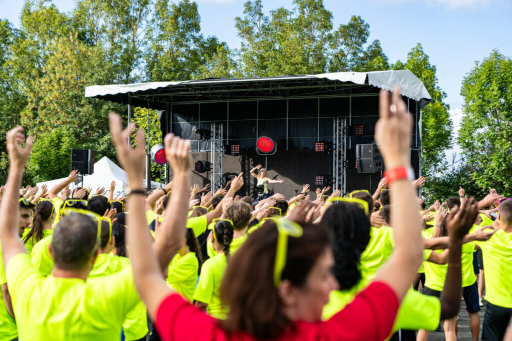 Groupe de personnes en tenue fluorescent, dansant et suivant une animatrice sur une scène en plein air.
