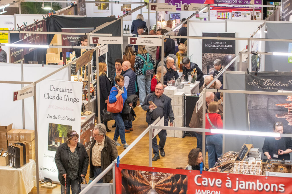 Salon où des visiteurs explorent des stands de produits locaux, notamment des vins et des spécialités gastronomiques.